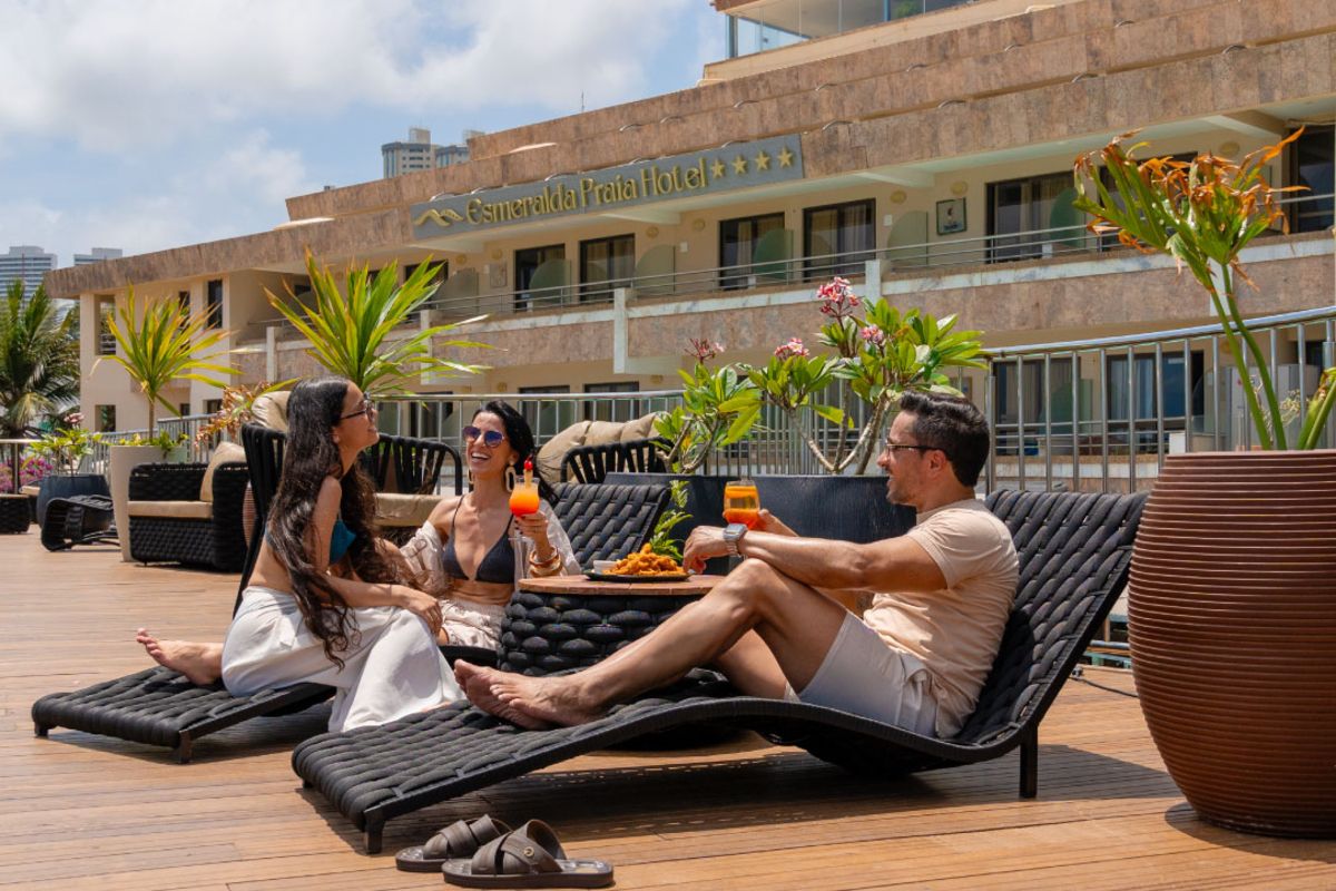 Hóspedes relaxam em espreguiçadeiras no deck do Esmeralda Praia Hotel com drinks e petiscos.